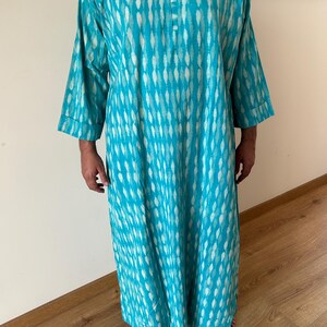 May include: A turquoise blue long-sleeved tunic with a white and turquoise pattern. The tunic has a round neckline and a button closure at the neck.