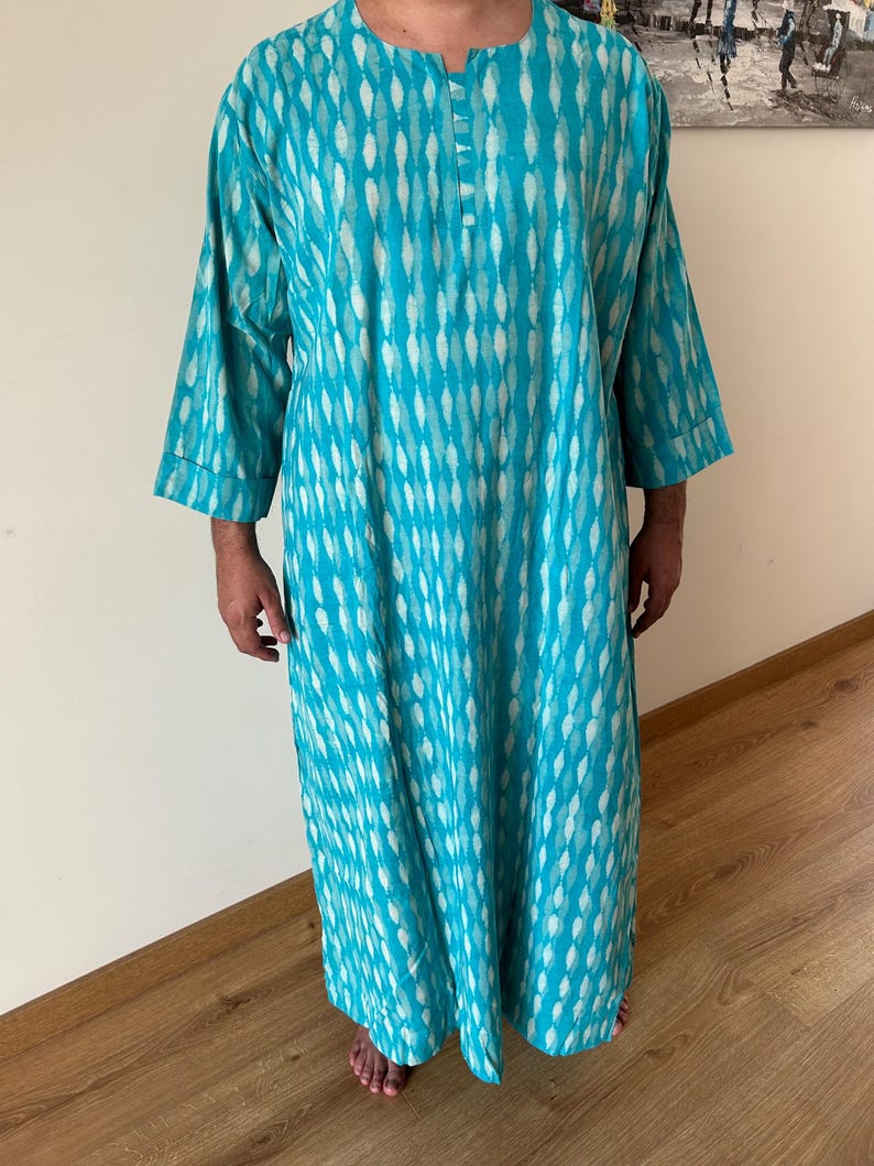 May include: A turquoise blue long-sleeved tunic with a white and turquoise pattern. The tunic has a round neckline and a button closure at the neck.