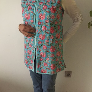 Könnte beinhalten: Eine lange, gesteppte Weste mit einem Blumendruck in Rosa- und Blautönen. Die Weste hat einen Stehkragen und einen Knopfverschluss.