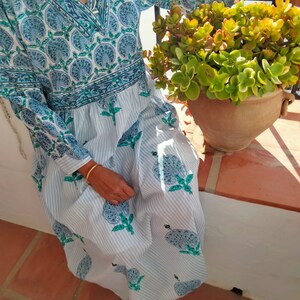 May include: A white cotton dress with blue and green floral print. The dress has a V-neck and long sleeves. The dress is worn by a person sitting on a patio.