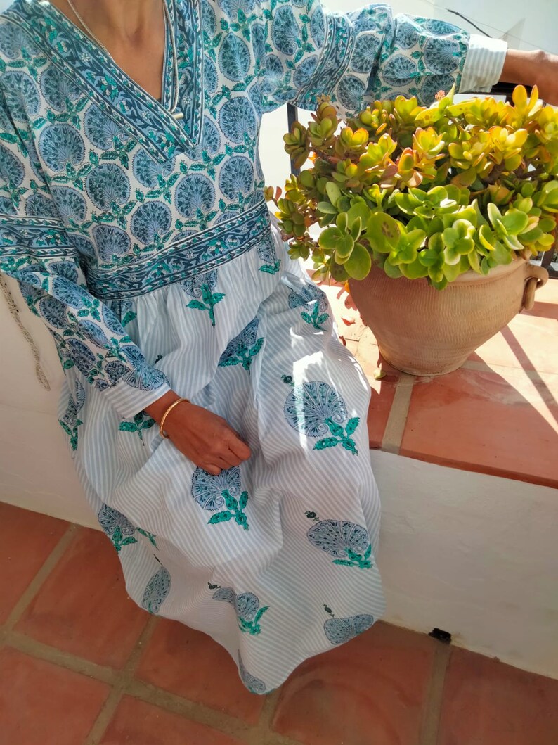 May include: A white cotton dress with blue and green floral print. The dress has a V-neck and long sleeves. The dress is worn by a person sitting on a patio.