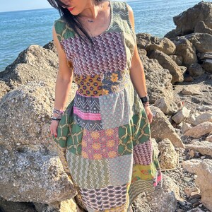 Pode incluir: Um vestido maxi sem mangas em patchwork com vários padrões e cores, incluindo verde, rosa e roxo. O vestido é usado com sandálias pretas. O fundo mostra uma costa rochosa e o mar. O vestido tem aproximadamente 150 cm de comprimento.
