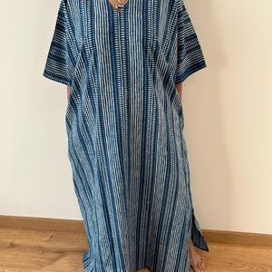 May include: A floor-length kaftan dress featuring a deep V-neckline and short sleeves. The garment is patterned with vertical stripes in shades of dark blue and white, incorporating a repeating dash-like texture throughout the fabric design.