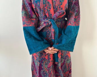 Puede incluir: Bata con estampado de cachemir en colores vibrantes, con borde y cinturón turquesa. La tela presenta una mezcla de tonos rojos, morados y turquesas. La bata es de tejido suave y texturizado, atada a la cintura.