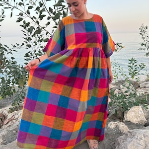 May include: A colorful, plaid maxi dress in shades of orange, pink, blue, and green. The dress has short sleeves and a round neckline. The model is wearing sandals and standing near the water.