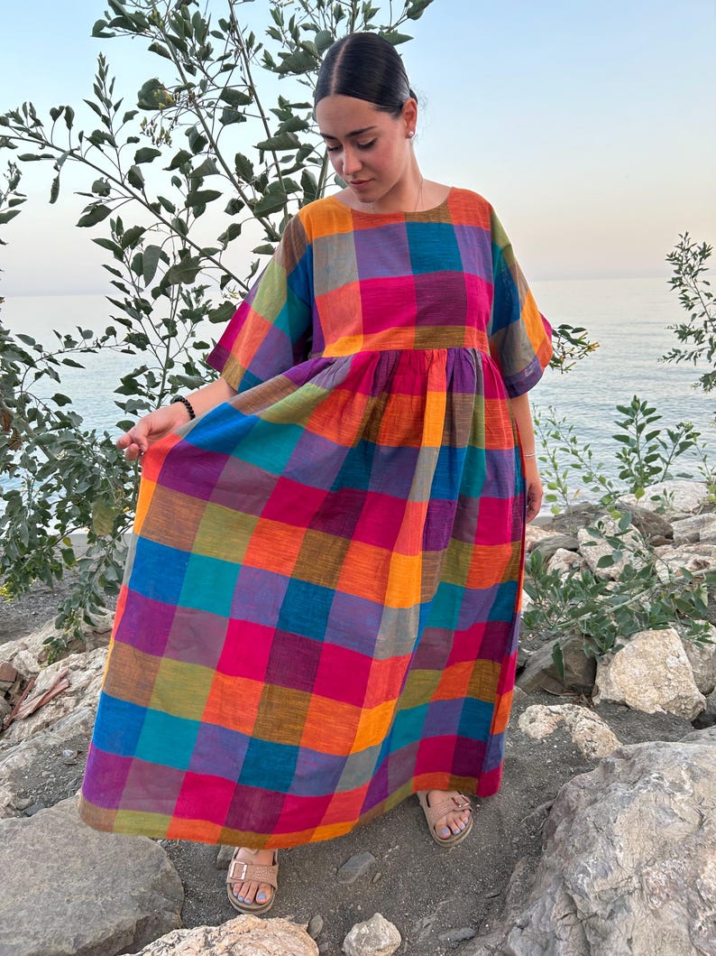 May include: A colorful, plaid maxi dress in shades of orange, pink, blue, and green. The dress has short sleeves and a round neckline. The model is wearing sandals and standing near the water.