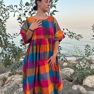 May include: A colorful, plaid maxi dress in shades of orange, pink, blue, purple, and green. The dress has short sleeves and a loose, flowing fit. The model is standing on rocks near the ocean.