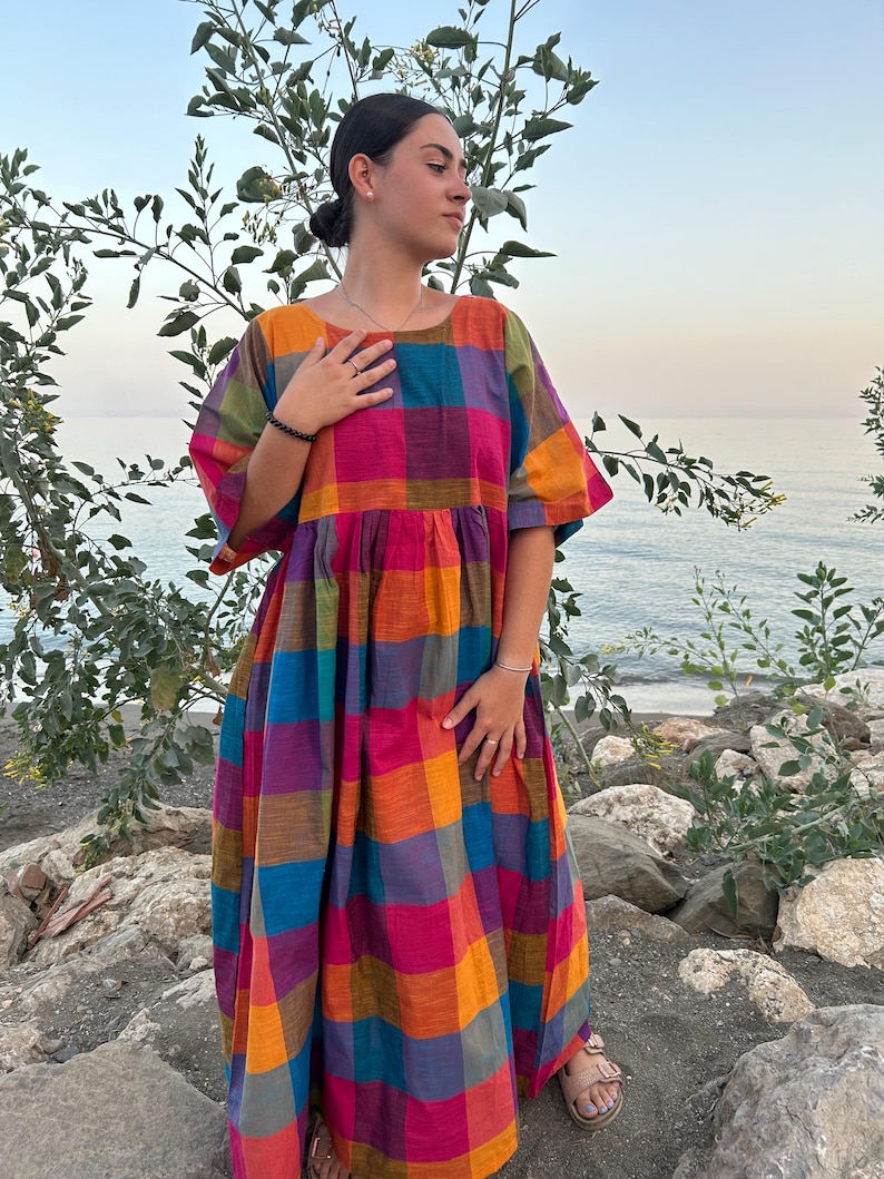 May include: A colorful, plaid maxi dress in shades of orange, pink, blue, purple, and green. The dress has short sleeves and a loose, flowing fit. The model is standing on rocks near the ocean.