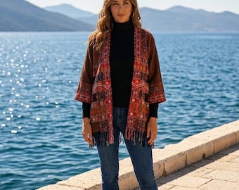 Puede incluir: Un kimono marrón abierto por delante con un patrón geométrico multicolor en tonos rojos, naranjas y azules, con mangas tres cuartos y un dobladillo con flecos, usado sobre un suéter de cuello alto negro y jeans azules.