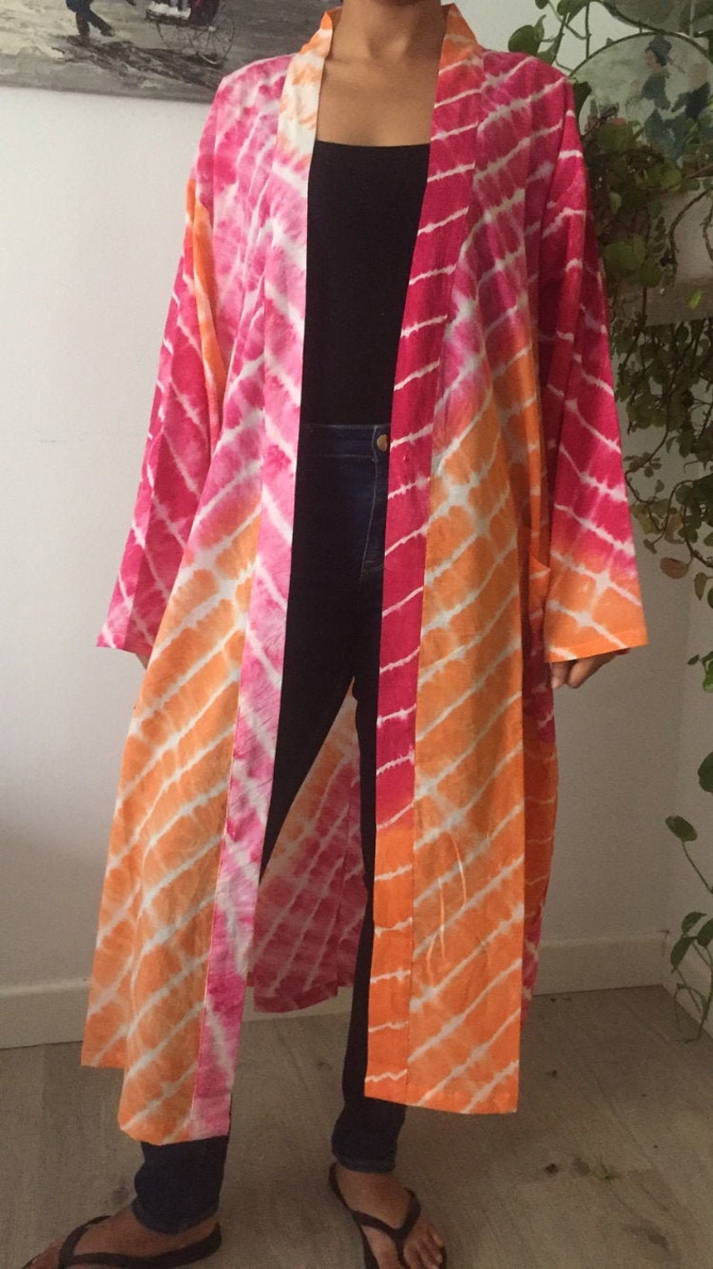 May include: A long, pink and orange tie-dye kimono with a white background. The kimono has a diagonal pattern and is open in the front. The person wearing the kimono is wearing black jeans and black sandals.