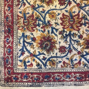 May include: A white cotton rug with a blue, red, and yellow floral pattern. The rug has a fringe border.