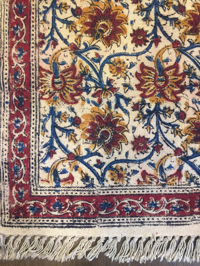 May include: A white cotton rug with a blue, red, and yellow floral pattern. The rug has a fringe border.