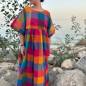 Può includere: Un abito a quadri colorato nei toni dell'arancione, rosa, blu e viola. L'abito ha maniche corte e una vestibilità ampia e fluida. L'abito è indossato all'aperto vicino al mare.