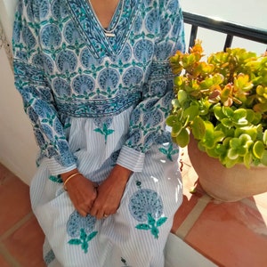 May include: A long-sleeved, white and blue dress with a floral print. The dress has a V-neckline and a gathered skirt. The dress is worn by a person sitting on a ledge.