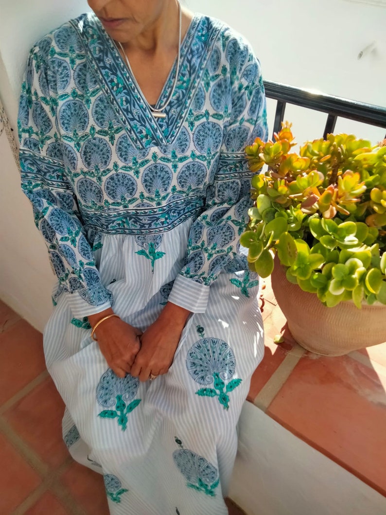 May include: A long-sleeved, white and blue dress with a floral print. The dress has a V-neckline and a gathered skirt. The dress is worn by a person sitting on a ledge.