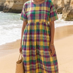 May include: A colorful plaid dress in yellow, green, purple, and pink. The dress has short sleeves and a gathered waist. A woven straw tote bag is held in the hand. The background shows a beach with sand and water.