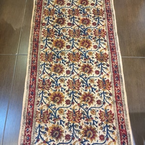 May include: A white cotton runner rug with a floral pattern in shades of red, blue, and yellow. The rug has a red border with a floral design and fringe on the ends.