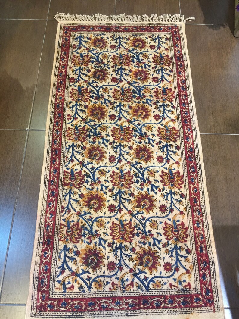 May include: A white cotton runner rug with a floral pattern in shades of red, blue, and yellow. The rug has a red border with a floral design and fringe on the ends.