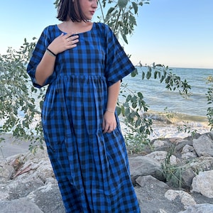 Op de afbeelding: Een blauwe en zwarte geruite maxi-jurk met een losse pasvorm en korte mouwen. De jurk is gefotografeerd op een persoon die op een rotsachtig strand staat.