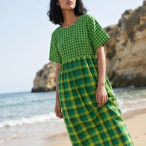 May include: A green and yellow plaid dress with short sleeves. The dress is knee-length and has a fitted bodice and a flared skirt. The model is standing on a sandy beach with the ocean in the background.