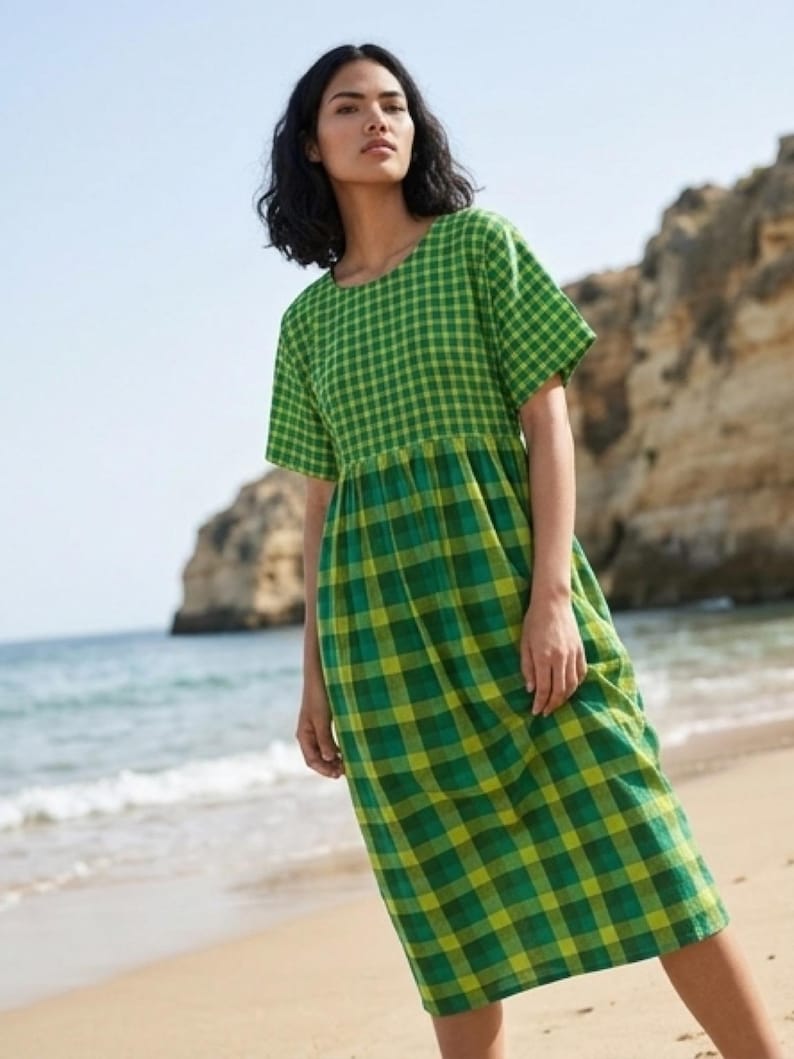 May include: A green and yellow plaid dress with short sleeves. The dress is knee-length and has a fitted bodice and a flared skirt. The model is standing on a sandy beach with the ocean in the background.