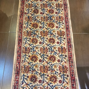 May include: A rectangular cream-colored rug with a floral pattern in shades of red, blue, and yellow. The rug has a red border with a blue and white pattern. The rug has fringe on the ends.