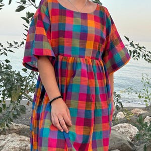 May include: A colorful plaid dress in shades of orange, pink, blue, and purple. The dress has short sleeves and a gathered waist. The background shows a beach scene with rocks and water. The dress is made of a lightweight fabric.