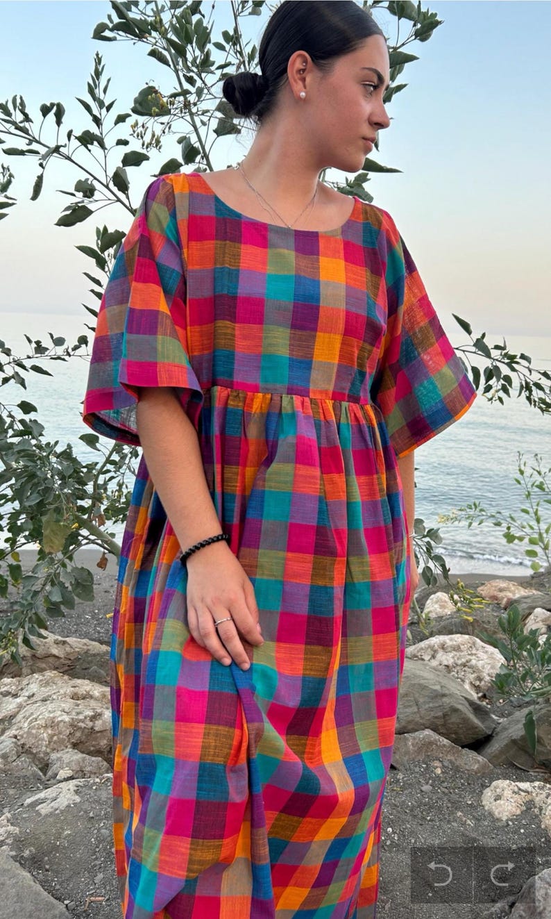 May include: A colorful plaid dress in shades of orange, pink, blue, and purple. The dress has short sleeves and a gathered waist. The background shows a beach scene with rocks and water. The dress is made of a lightweight fabric.