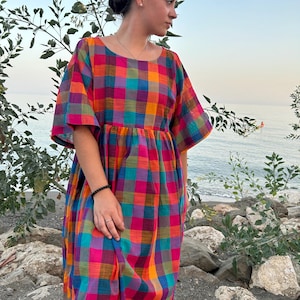 Può includere: Una donna indossa un abito maxi a quadri colorati con maniche corte. L'abito ha una vestibilità ampia e una vita arricciata. La donna è in piedi su una spiaggia rocciosa con l'oceano sullo sfondo.