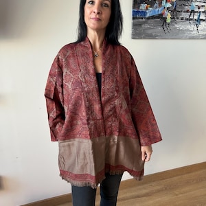 Puede incluir: Chaqueta estilo kimono burdeos y marrón con estampado de cachemira y dobladillo con flecos. La chaqueta está abierta por delante y se lleva sobre un top negro. El diseño de la chaqueta presenta un patrón detallado y repetitivo.