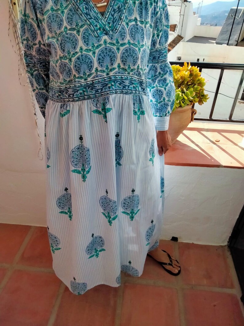 May include: A long white dress with blue and green floral print. The dress has a V-neck and long sleeves. The skirt is gathered at the waist and has a striped pattern.