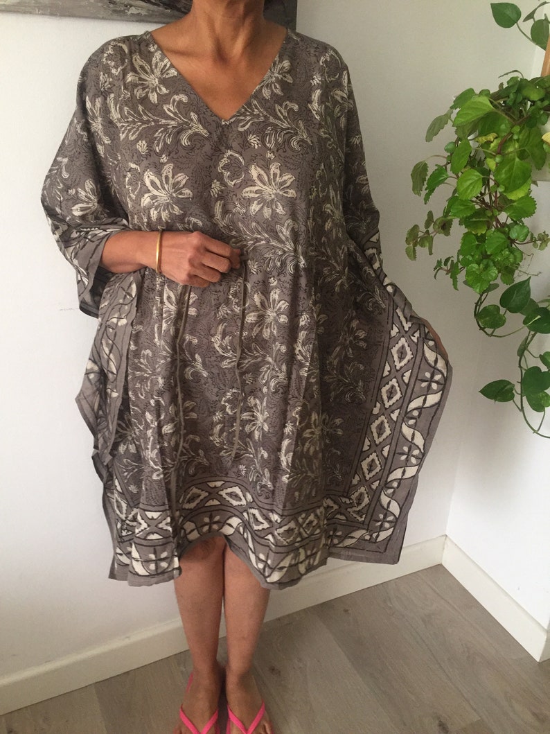 May include: A gray and white floral print kaftan with a geometric border. The kaftan has a V-neck and 3/4 sleeves.