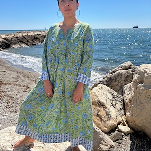 Könnte beinhalten: Eine Frau trägt ein langes, grünes und blaues Blumenkleid mit V-Ausschnitt und langen Ärmeln. Sie steht auf einem felsigen Strand mit dem Meer im Hintergrund.