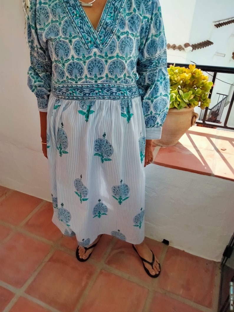 May include: A long, white dress with blue and green floral print. The dress has a V-neckline and a gathered waist. The skirt is striped and has a flowing silhouette.