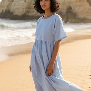 May include: A light blue, short-sleeved dress with a small white checkered pattern. The dress has a gathered waist and falls to mid-calf. The model is standing on a sandy beach with the ocean in the background.