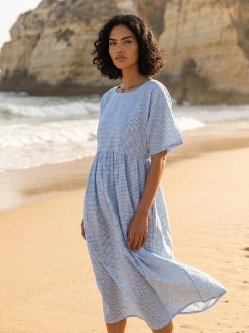May include: A light blue, short-sleeved dress with a small white checkered pattern. The dress has a gathered waist and falls to mid-calf. The model is standing on a sandy beach with the ocean in the background.