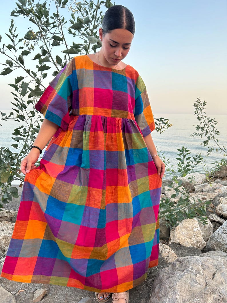 May include: A colorful, plaid-patterned dress in shades of orange, pink, purple, green, and blue. The dress features a round neckline, short sleeves, and a gathered waist. The long, flowing skirt falls to the ankles. The dress is made of a lightweight fabric, perfect for warm weather.