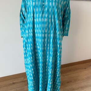 May include: A turquoise blue long-sleeved tunic with a white and turquoise pattern. The tunic has a round neckline and a button closure at the neck.