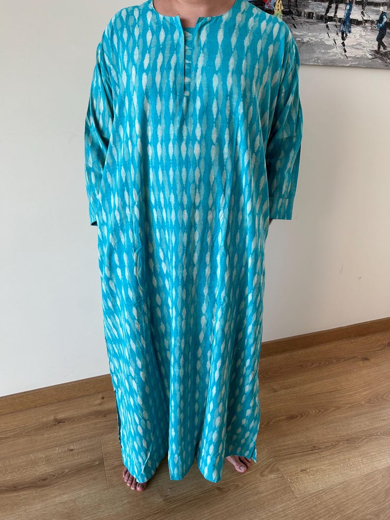 May include: A turquoise blue long-sleeved tunic with a white and turquoise pattern. The tunic has a round neckline and a button closure at the neck.