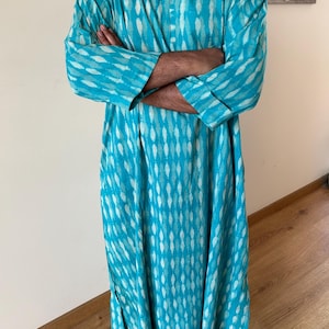 May include: A long, turquoise blue tunic with a white geometric pattern. The tunic has a button-up neckline and long sleeves.