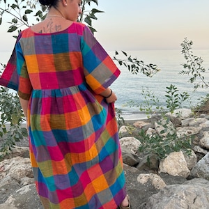 May include: A colorful, long, plaid dress in shades of pink, orange, blue, and green. The dress has short sleeves and a gathered waist. The person is standing on rocks near the water.