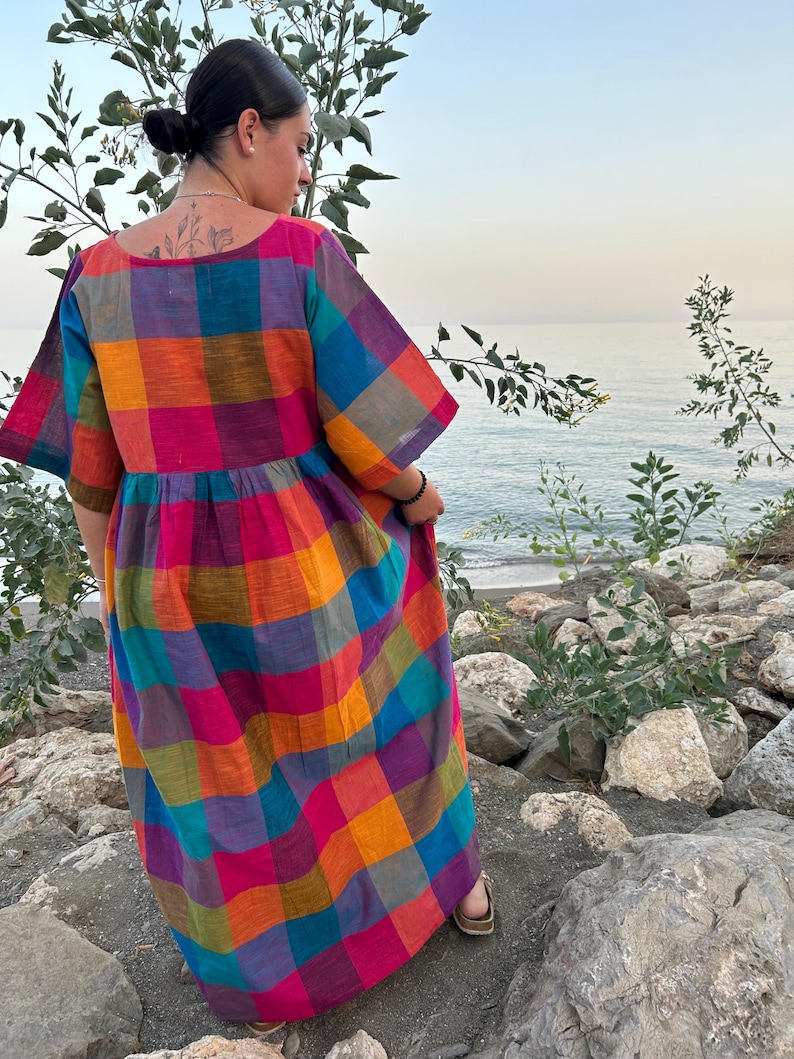 May include: A colorful, long, plaid dress in shades of pink, orange, blue, and green. The dress has short sleeves and a gathered waist. The person is standing on rocks near the water.