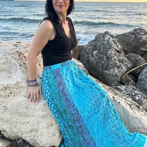 Puede incluir: Un top negro con ribete de encaje y una falda larga y fluida de color turquesa con un diseño estampado. La falda presenta una mezcla de tonos azules, morados y verdes. La persona lleva gafas de sol y pulseras.