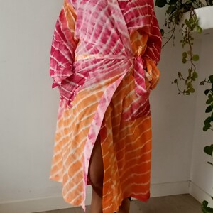 May include: A pink and orange tie-dye robe with a long sleeve and a wrap closure. The robe has a side slit and is made of a soft, lightweight fabric.