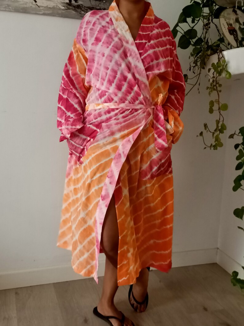 May include: A pink and orange tie-dye robe with a long sleeve and a wrap closure. The robe has a side slit and is made of a soft, lightweight fabric.