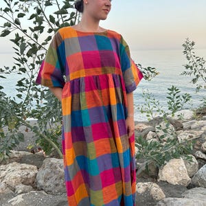 May include: A colorful, long, plaid dress in shades of pink, orange, purple, blue, and green. The dress has short sleeves and a loose, flowing fit. The model is standing on a rocky beach with the ocean in the background.