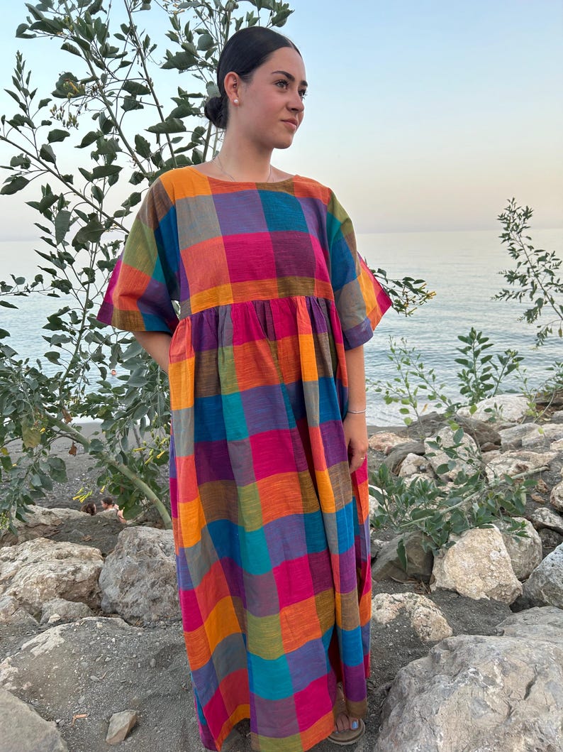 May include: A colorful, long, plaid dress in shades of pink, orange, purple, blue, and green. The dress has short sleeves and a loose, flowing fit. The model is standing on a rocky beach with the ocean in the background.
