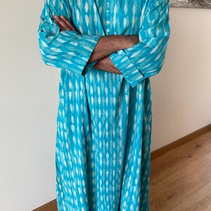May include: A long, turquoise blue tunic with a white geometric pattern. The tunic has a V-neck and long sleeves with button cuffs.