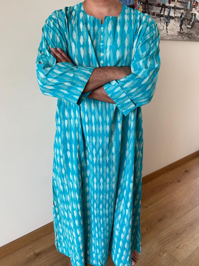 May include: A long, turquoise blue tunic with a white geometric pattern. The tunic has a V-neck and long sleeves with button cuffs.