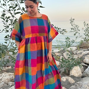 May include: A colorful, patterned dress with a square neckline and short sleeves. The dress features a vibrant plaid design in shades of orange, pink, purple, blue, and green. The dress is worn outdoors.